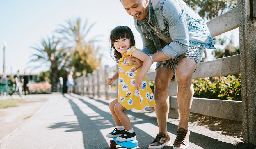 Man helping little girl in yellow dress ride a skateboard
