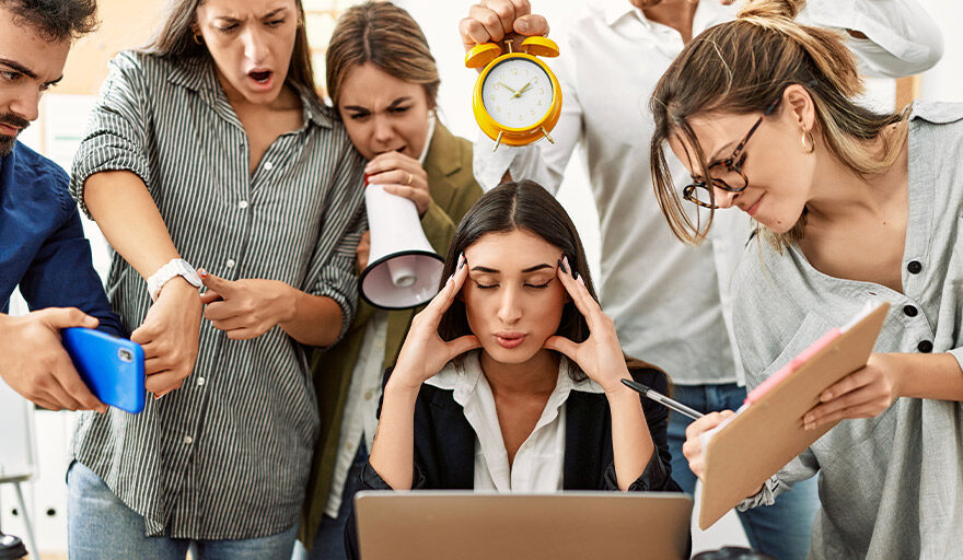 Woman at a desk with hands on her head surrounded by people asking her for things.