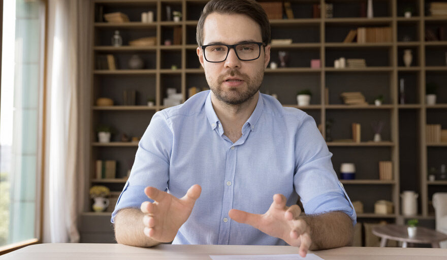 Man in business casual clothes siting at a desk having a conversation with the camera