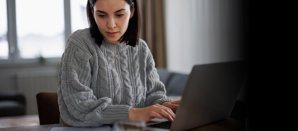 Woman wearing a gray sweater working on a laptop at home.