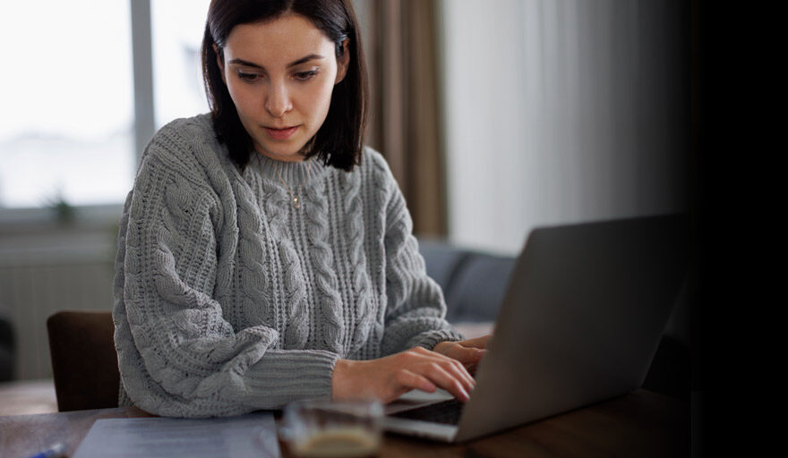 Woman wearing a gray sweater working on a laptop at home.